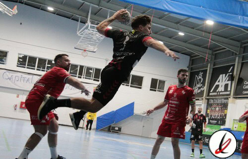 Saltador en acción durante un partido de balonmano en un gimnasio cubierto, con jugadores en uniforme rojo y fondo con anuncios y estructura deportiva.