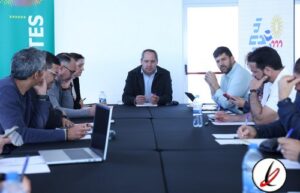 Reunión de un grupo de personas en una mesa de conferencia, con un hombre en el centro hablando y otros tomando notas o usando dispositivos electrónicos.