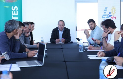 Reunión de un grupo de personas en una mesa de conferencia, con un hombre en el centro hablando y otros tomando notas o usando dispositivos electrónicos.