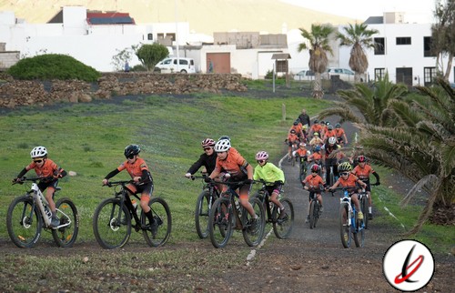 Andar en bicicleta por un sendero de tierra con un grupo de ciclistas, algunos con cascos y ropa deportiva, en un entorno natural con palmeras y casas blancas al fondo.
