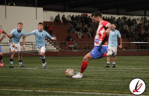Disparo de un jugador de fútbol en un partido en un campo cubierto, con defensores en posición de bloqueo y público en las gradas.