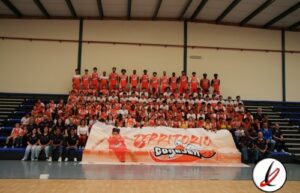 Jugadores de baloncesto en un equipo, posando en un gimnasio con un banner y espectadores en las gradas.