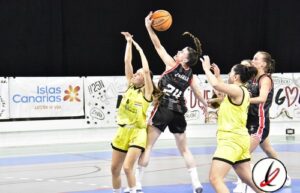 Jugadora de baloncesto en acción durante un partido, saltando para encestar mientras es defendida por oponentes en la cancha cubierta.