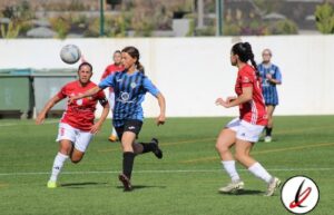 Jugadoras de fútbol femenino en acción durante un partido en un campo al aire libre, con una jugadora en azul controlando el balón y otras en rojo defendiendo.