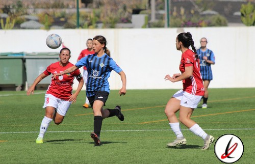 Jugadoras de fútbol femenino en acción durante un partido en un campo al aire libre, con una jugadora en azul controlando el balón y otras en rojo defendiendo.