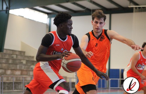 Baloncestistas en acción durante un partido en una cancha cubierta, con jugadores en uniforme naranja y negro, y uno en uniforme rojo y blanco, en un ambiente deportivo interior.