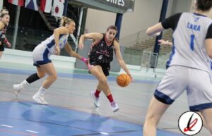 Jugadora de baloncesto en acción en una cancha cubierta, intentando avanzar con el balón mientras es defendida por dos oponentes.