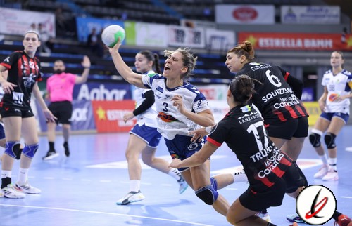 Balón de balonmano en juego durante un partido femenino en una pista cubierta. Jugadoras en acción, algunas intentando bloquear y otras lanzando, con público y anuncios en el fondo.