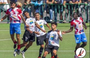 Jugadores de fútbol en acción durante un partido en un campo de césped, con público en las gradas al fondo.