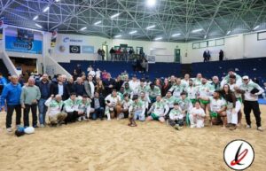 Jugadores de rugby en un campo de arena en un estadio cubierto, posando para la foto con sus entrenadores y seguidores.