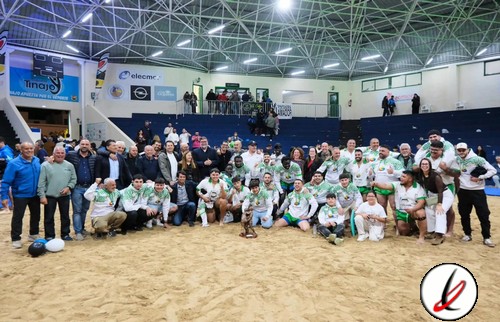 Jugadores de rugby en un campo de arena en un estadio cubierto, posando para la foto con sus entrenadores y seguidores.
