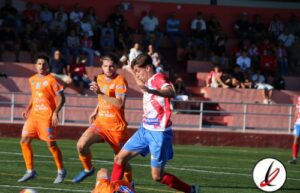 Jugadores de fútbol en acción durante un partido en un estadio al aire libre con espectadores en las gradas.