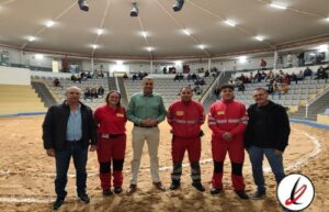 Grupo de cinco personas en un estadio cubierto, con tres en uniforme rojo y dos en ropa casual, en el centro del campo de arena.