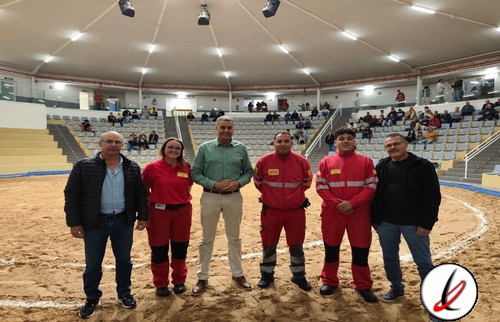Grupo de cinco personas en un estadio cubierto, con tres en uniforme rojo y dos en ropa casual, en el centro del campo de arena.