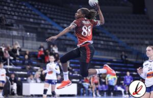 Atleta femenina en acción durante un partido de balonmano en el pabellón deportivo de Lanzarote. Jugadora saltando para lanzar o defender en un encuentro competitivo, con público y otras jugadoras en el fondo.