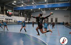 Una jugadora de balonmano en acción, saltando para lanzar el balón en un partido en un pabellón cubierto con espectadores y anuncios publicitarios en las paredes.