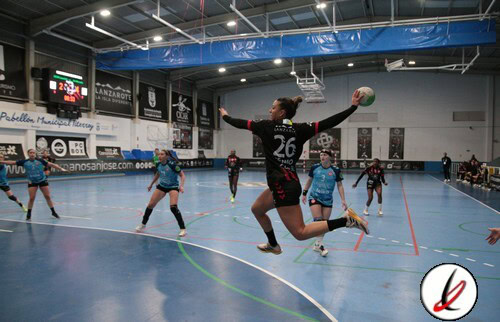 Una jugadora de balonmano en acción, saltando para lanzar el balón en un partido en un pabellón cubierto con espectadores y anuncios publicitarios en las paredes.