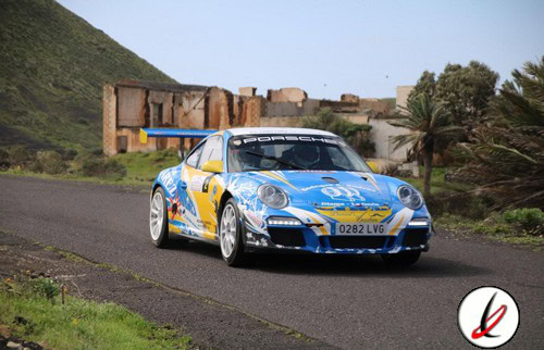 Porsche de rally en una carretera de Lanzarote, con paisaje volcánico y construcciones tradicionales en el fondo.