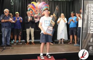 Joven en el escenario recibiendo un premio en un evento cultural, rodeado de personas que aplauden y observan la ceremonia, con un fondo colorido y decorativo en un espacio cerrado.