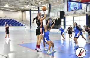 Baloncesto femenino en pista cubierta, jugadora en acción lanzando a canasta durante un partido en un gimnasio moderno con espectadores en las gradas.