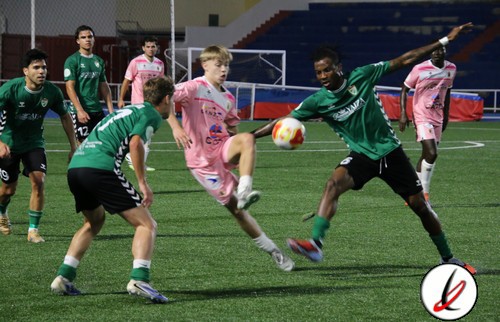 Jugadores de fútbol en acción durante un partido en un estadio cubierto, con jugadores en camisetas verdes y rosas, en movimiento y luchando por el balón.