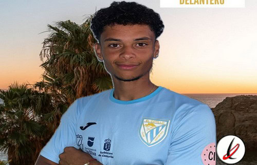 Jugador de fútbol joven con camiseta azul del equipo Lanzarote Deportivo, en un entorno al atardecer con palmeras y rocas en el fondo.