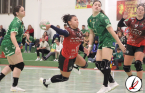 Jugadoras de balonmano en acción durante un partido en un gimnasio, con público en las gradas y cartelera en el fondo.