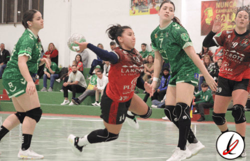 Jugadoras de balonmano en acción durante un partido en un gimnasio, con público en las gradas y cartelera en el fondo.