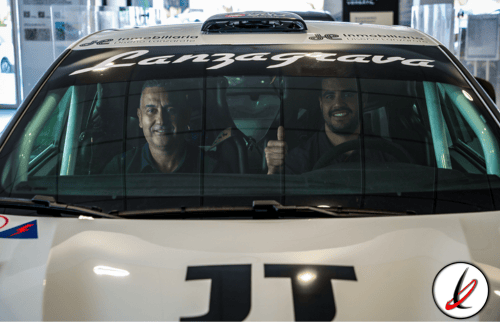 Línea 1 (ALT tag): Conductores sonriendo en un coche de carreras con logotipo Lanzarote en el parabrisas, en un entorno de exposición o evento automovilístico.
