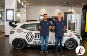 Dos hombres de pie junto a un coche de carreras en un concesionario, con fondo de vitrinas y pantallas, en un entorno moderno y bien iluminado.