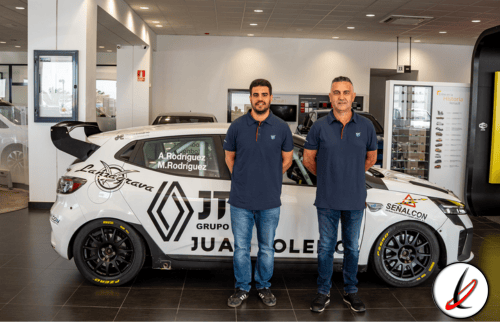 Dos hombres de pie junto a un coche de carreras en un concesionario, con fondo de vitrinas y pantallas, en un entorno moderno y bien iluminado.