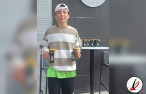 Joven con gorra blanca, camiseta a rayas beige y blanca, pantalones cortos verdes y negros, sosteniendo dos trofeos en un entorno moderno.