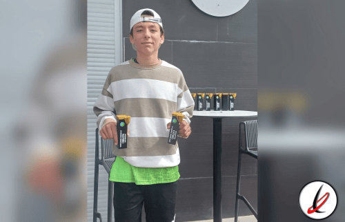 Joven con gorra blanca, camiseta a rayas beige y blanca, pantalones cortos verdes y negros, sosteniendo dos trofeos en un entorno moderno.
