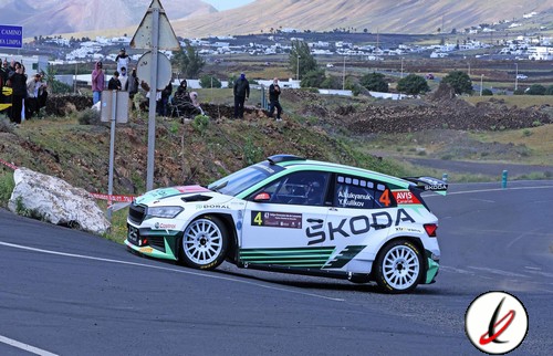 Coche de rally de color blanco con detalles verdes y negros en una curva de carretera en Lanzarote, con espectadores observando en el fondo y un paisaje volcánico.