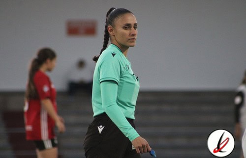 Árbitra de fútbol en un campo de interior, vestida con uniforme verde y pantalones negros, observando el juego con atención.