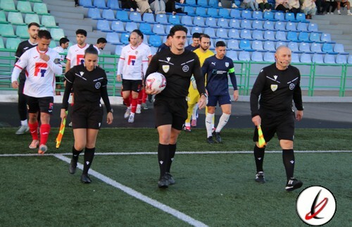 Árbitros y jugadores de fútbol en el campo antes del inicio del partido en un estadio con gradas vacías.