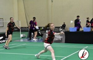 Jugadoras de bádminton en acción en una cancha cubierta, con una de ellas en movimiento preparándose para golpear el volante.