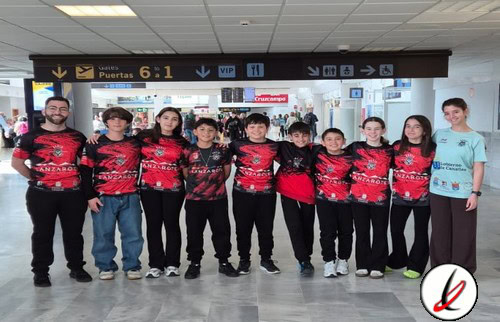 Grupo de niños y jóvenes en aeropuerto, algunos con camisetas de deporte, en fila, con un adulto a la izquierda y una mujer a la derecha, fondo de señalización de aeropuerto.
