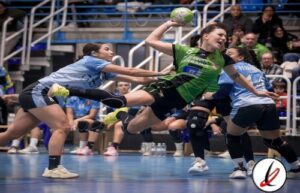 Jugadora de balonmano en acción, lanzando el balón en un partido femenino en un pabellón deportivo.