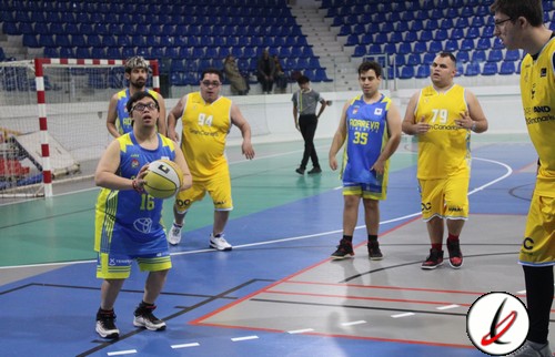 Balón de baloncesto en manos de un jugador en una cancha cubierta. Jugadores en uniforme amarillo y azul, algunos observando, en un partido de baloncesto en interiores con gradas vacías.