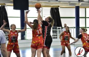 Taga de baloncesto femenino en acción durante un partido en una cancha cubierta, con jugadoras en movimiento y una jugadora en el aire intentando encestar.