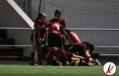 Jugadoras de fútbol femenino celebrando un gol en el campo, con algunas en el suelo y otras de pie, en un estadio con fondo de malla y paredes blancas.