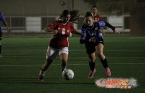 Línea 1 (ALT tag): Jugadoras de fútbol femenino en acción durante un partido nocturno en un campo de césped artificial.