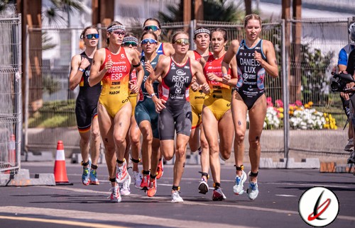 Grupo de atletas femeninas participando en una carrera en una calle de Lanzarote, con fondo de vallas y flores, en un evento deportivo al aire libre.