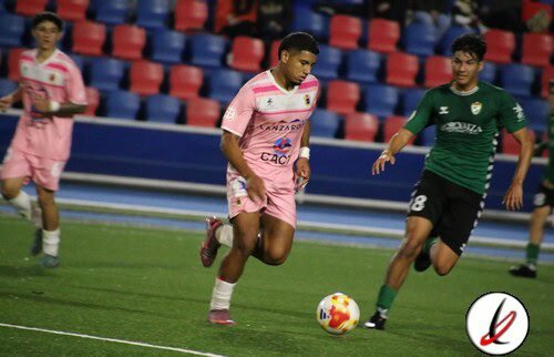 Jugador en camiseta rosa controlando el balón en un partido de fútbol en un estadio con gradas rojas y azules.