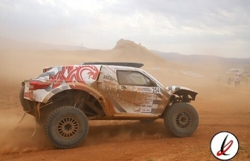 Vehículo de rally raid en movimiento en un terreno árido con polvo en suspensión y un paisaje montañoso al fondo.