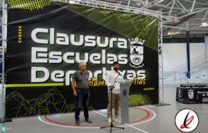 Dos hombres en un acto de clausura en un gimnasio, uno con camiseta gris y el otro con camisa blanca, frente a un gran cartel que dice "Clausura Escuelas Deportivas".