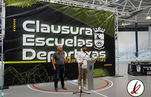Dos hombres en un acto de clausura en un gimnasio, uno con camiseta gris y el otro con camisa blanca, frente a un gran cartel que dice "Clausura Escuelas Deportivas".