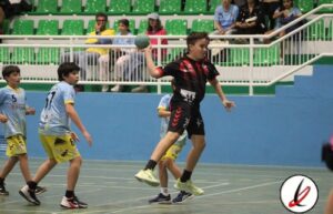 Balonmano infantil en un pabellón deportivo con jugadores en acción y espectadores en las gradas.