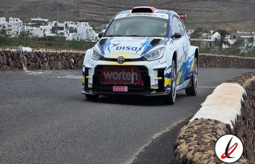 Coche de rally en una curva de carretera en Lanzarote, con montañas y casas blancas al fondo.
